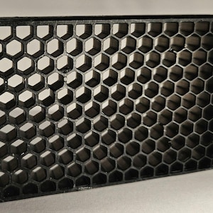 Puede incluir: Una rejilla negra rectangular con forma de panal de abejas y un patrón hexagonal repetitivo. La rejilla está hecha de un material oscuro y tiene una apariencia tridimensional debido a la profundidad de las celdas. El diseño es un patrón geométrico.