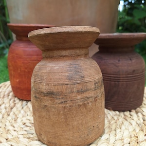 Könnte beinhalten: Drei handgefertigte Holzvasen in verschiedenen Braun- und Orangetönen stehen auf einer gewebten Matte. Die Vasen haben einen abgerundeten Körper und eine ausgestellte Oberseite. Der Hintergrund zeigt einen großen Terrakottatopf und grünes Laub.
