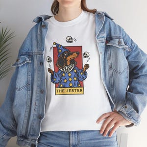 Może przedstawiać: Biały t-shirt z grafiką jamnika przebranego za błazna w niebieskiej szacie i czapce w gwiazdki, żonglującego trzema czaszkami. Wzór jest ujęty w czerwoną ramkę z żółtym napisem THE JESTER umieszczonym w dolnej części prostokątnego nadruku.