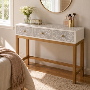 May include: A rectangular console table with three drawers featuring a white and gray floral pattern and gold-toned knobs. The table has a white top surface and four light brown wooden legs connected by a lower support frame for stability.