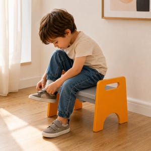 Op de afbeelding: Een kleine, lage kruk met een lichtgrijze rechthoekige zitting en twee feloranje zijpanelen met geïntegreerde handgrepen. De kruk is stevig geconstrueerd en biedt een stabiele basis om op te zitten.