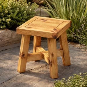 Puede incluir: Un taburete cuadrado de madera con un acabado natural en color marrón claro, que presenta una superficie superior plana, cuatro patas robustas y una estructura de soporte en cruz que conecta las patas cerca de la base, con vetas y nudos visibles.