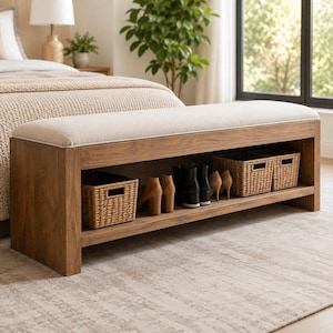 Può includere: Una panca rettangolare in legno con un cuscino di seduta imbottito color beige chiaro. La base presenta un ripiano aperto contenente un cesto intrecciato a sinistra, un paio di stivali con tacco color cuoio, un paio di stivali neri e due cesti intrecciati.