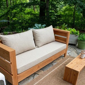 May include: A wooden outdoor sofa with two beige cushions and pillows. The sofa is on a brown rug, with a wooden coffee table to the right. The background features lush green trees and plants, creating a natural setting.