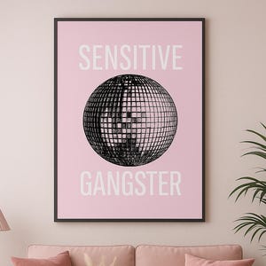 May include: A framed art print with the words "SENSITIVE" and "GANGSTER" above and below a black and white disco ball graphic. The print hangs above a pink sofa with matching pillows and a white coffee table.