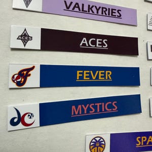 May include: A close-up of several rectangular team name badges. The badges feature team names like "VALKYRIES", "ACES", "FEVER", and "MYSTICS" in various colors. Each badge has a team logo on the left side.