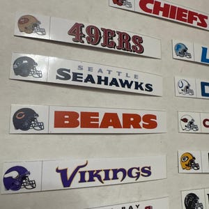 May include: Rectangular white stickers featuring NFL team logos and names. The stickers include the "49ERS", "SEAHAWKS", "BEARS", "VIKINGS", "CHIEFS", and "BUCCANEERS" logos. Each sticker has a team helmet graphic.