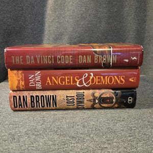 May include: Three books stacked, featuring titles by Dan Brown. The top book is "The Da Vinci Code". The middle book is "Angels & Demons". The bottom book is "The Lost Symbol". All books have a red and brown color scheme.