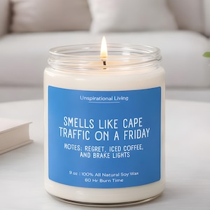May include: A lit candle in a glass jar with a blue label. The label reads "Smells Like Cape Traffic on a Friday" with notes of regret, iced coffee, and brake lights. The candle is 9 oz and made of 100% all-natural soy wax.