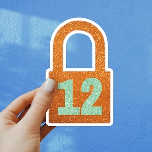 May include: An orange glitter sticker in the shape of a padlock, with the number 12 in teal. The sticker is held by a hand against a blue background. The sticker has a white border.