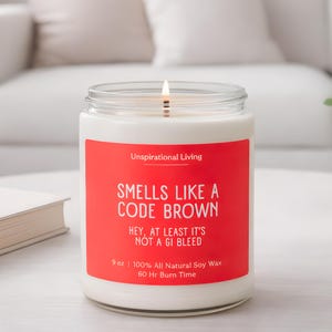 Peut inclure: Une bougie blanche allumée dans un pot en verre avec une étiquette rouge. L'étiquette indique "SMELLS LIKE A CODE BROWN" et "HEY, AT LEAST IT'S NOT A GI BLEED." La bougie est de 255 g et faite de cire de soja 100% naturelle.