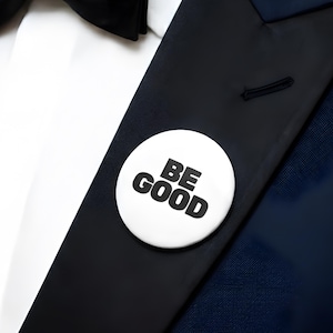 May include: A white button with the words "BE GOOD" in bold black letters is pinned to a black lapel. The lapel is part of a dark blue suit jacket, with a white dress shirt and black bow tie visible.