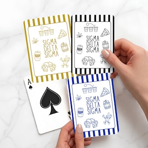 Op de afbeelding: Drie speelkaarten met gestreepte randen in goud, zwart en blauw. Elke kaart bevat de tekst Sigma Delta Sigma omringd door lijntekeningen van beer pong, pizza, een bierblikje, een vat, bierpullen en een cannabisblad.