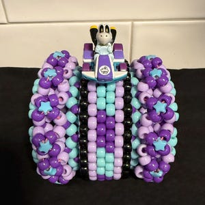 Könnte beinhalten: Ein handgefertigtes Kandi-Armband mit einem Spielzeug-Rennwagen und einer Charakterfigur. Das Armband besteht aus lila, hellblauen und schwarzen Perlen mit sternförmigen Akzenten. Der Rennwagen ist lila und weiß.