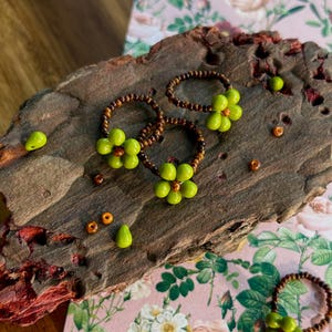 May include: Handmade beaded rings with a floral design. The rings feature a brown beaded band with a cluster of green beads in the shape of a flower. Additional loose beads in green and orange are scattered around the rings on a wooden surface.