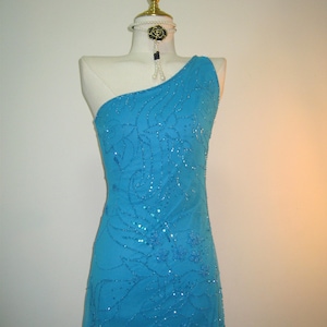 以下が含まれることがあります： トルコ石色のワンショルダーイブニングドレスがマネキンに飾られています。ドレスはスパンコールと刺繍の花柄で装飾されています。黒いバラのペンダントが付いた真珠のネックレスがマネキンの首を飾っています。