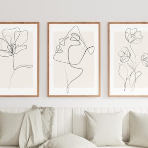 以下が含まれることがあります： ミニマリストの線画が描かれた、額装されたアートプリント3点。プリントには、花、顔、その他の花が描かれており、すべてベージュの背景に黒い線で描かれています。フレームは木製です。アートワークは、ベージュのクッションが付いた白いソファの上に展示されています。