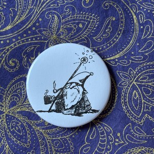 May include: A white circular badge with a cartoon illustration of a badger dressed as a wizard, holding a staff with sparkles and smoking a pipe. The background is a dark blue fabric with a gold paisley pattern.