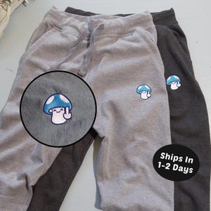 May include: Two pairs of gray and charcoal sweatpants with a mushroom graphic embroidered on the left leg. The mushroom has a blue and white cap and is giving the middle finger. The text "Ships In 1-2 Days" is on a black circle.