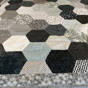 May include: A handmade quilt featuring a geometric pattern of hexagonal patches in various shades of gray, black, and white. The quilt showcases a variety of textures and patterns, including polka dots, stripes, and floral designs. The border has a gray and white leaf pattern.