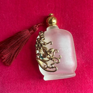 May include: Frosted glass perfume bottle with a gold-colored filigree design and a red tassel. The bottle has a gold-colored cap and a rectangular shape. The filigree features grapes and leaves.