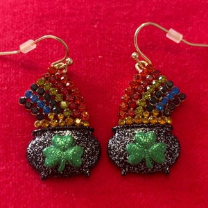 May include: A pair of gold-tone earrings featuring a black pot with a green shamrock and a rainbow of colorful gems. The earrings have a hook closure and are set against a red background.