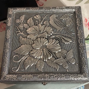 May include: A square silver-toned metal box featuring an embossed lid with a detailed design of two large hibiscus flowers, two butterflies, and a bird perched on a branch. The border is decorated with a repeating floral pattern and beaded edges.