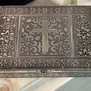 May include: A rectangular silver-toned metal box featuring an intricate embossed floral pattern along the borders and a central panel displaying a prominent cross symbol surrounded by detailed scrollwork and leaf motifs with a small front latch.