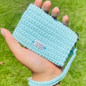 May include: A light blue rectangular crochet pouch featuring a textured stitch pattern and a matching wrist strap. A small white rectangular tag is attached to the front, clearly displaying the black printed text, Handmade with love.