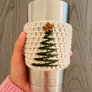 May include: A cream-colored, crocheted coffee cup cozy with a green Christmas tree design. A small wooden snowflake is attached at the top. The cozy is wrapped around a silver Yeti tumbler.