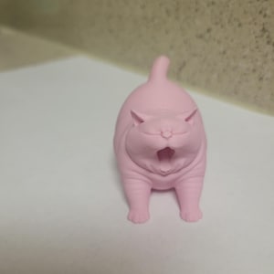 May include: A pink cat ornament with an open mouth, possibly yawning. The cat is standing upright with its tail raised. The ornament is made of a solid material and has a smooth texture. The cat's features are clearly defined.