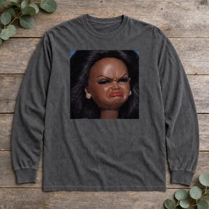 Op de afbeelding: Een antracietgrijs t-shirt met lange mouwen en een gecentreerde vierkante grafische print van een pop met donker haar, gefronste wenkbrauwen en een pruilende uitdrukking. De pop heeft opvallende rode lippen en lichte oorbellen op een blauwe achtergrond.