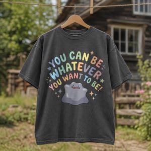 May include: A charcoal grey short-sleeve t-shirt featuring a graphic of a purple blob character and the text "YOU CAN BE WHATEVER YOU WANT TO BE!" in pastel-coloured letters, accented with small star shapes surrounding the central design.