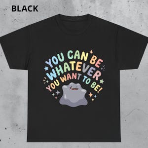 Op de afbeelding: Een zwart T-shirt met korte mouwen en een centrale afbeelding met de tekst "YOU CAN BE WHATEVER YOU WANT TO BE!" in pastelkleuren. Onder de tekst staat een paars, amorf, klodderachtig figuurtje met een eenvoudig gezicht, omringd door sterretjes.