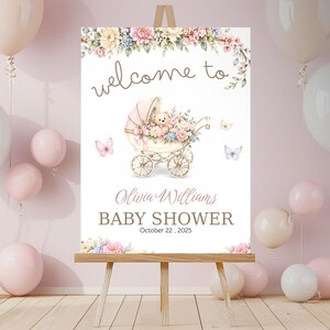 May include: A baby shower welcome sign on an easel. The sign features floral illustrations, butterflies, and a baby carriage. Text reads "welcome to" and "Baby Shower" with a date and name.