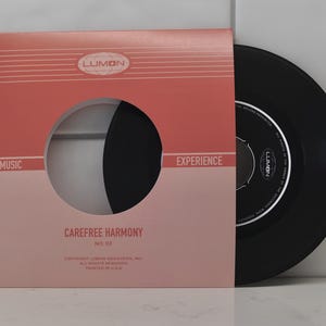 May include: A vintage 45 RPM record in a pink and red sleeve. The record is black and partially visible through a circular cutout. The sleeve has the text "LUMON", "MUSIC EXPERIENCE", and "CAREFREE HARMONY".