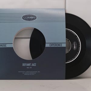 May include: A black vinyl record in a blue and grey sleeve. The sleeve has the text "LUMON", "MUSIC", "EXPERIENCE", and "DEFIANT JAZZ". The record label is visible, with the "LUMON" logo and text. A vintage music item.