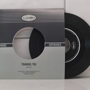 May include: A vintage 45 RPM vinyl record and sleeve. The sleeve is grey and black with the text "LUMON" and "TRANQUIL TIKI". The record is black and partially visible. The sleeve has a circular cutout.