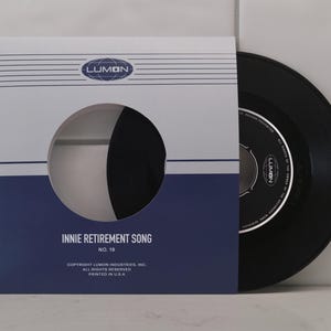 May include: A black vinyl record in a white and navy blue sleeve. The sleeve has a circular cut-out showing the record. Text on the sleeve reads "INNIE RETIREMENT SONG" and "LUMON".