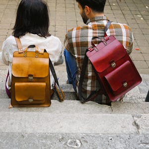 May include: Two leather backpacks, one tan and one burgundy, are on concrete steps. The tan backpack has a front pocket and a flap closure. The burgundy backpack has a front pocket and a buckle closure. Both have shoulder straps.