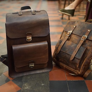 May include: Two brown leather backpacks. One is a tall, rectangular design with a top handle and a front pocket. The other is a more traditional backpack style with adjustable straps and buckle closures. Both have a vintage, distressed appearance.