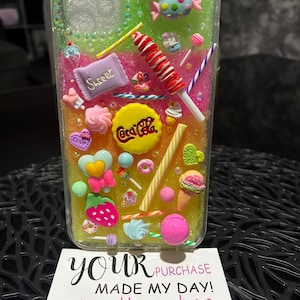 May include: A clear phone case adorned with colourful candy decorations, such as a lollipop, a Coca-Cola logo, and various sweets. The case has a glittery background and includes the text "YOUR PURCHASE MADE MY DAY! I hope this makes YOUR DAY!"