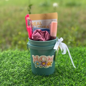 May include: A teal bucket filled with gifts, including a pink pen, a package with the text "REAL ATEA", a pink scrunchie, and a lipstick. The bucket has a "MOM" sticker with floral accents and a white ribbon tied to the handle.
