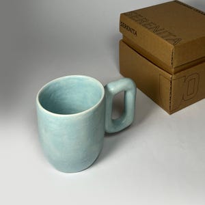 Könnte beinhalten: Hellblaue Keramiktasse mit einem einzigartigen quadratischen Griff. Die Tasse hat eine glatte, matte Oberfläche und einen abgerundeten Boden. Ein brauner Pappkarton mit dem Wort "SERENITA" befindet sich im Hintergrund.