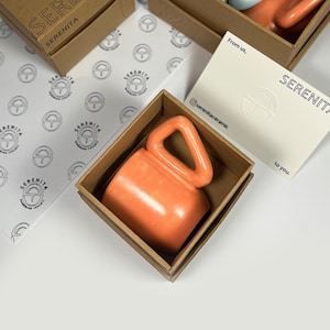 May include: An orange ceramic mug with a unique handle design, presented in a brown box. The box is labeled "SERENITA". Other mugs in blue and orange are visible in the background. A card with the text "From us, SERENITA" is also present.