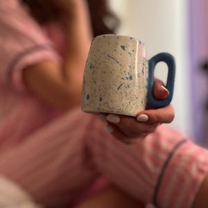 Puede incluir: Una taza de cerámica con un acabado beige moteado y un asa y salpicaduras azules. La taza se sostiene en la mano de una persona, con un fondo borroso de pijama rosa.