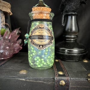 May include: A clear glass apothecary jar filled with small green spheres. The jar is sealed with a cork stopper topped by a small frog figurine. A label reads THE GOLDEN TOAD SINCE 1648 ENCHANTED FROG EGGS APOTHECARY. A black cord with two beads is tied around the neck.