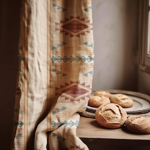 Peut inclure: Un textile beige avec un motif géométrique de losanges et de triangles dans des tons de rouge, turquoise et orange. Le textile est drapé sur une surface en bois, à côté d'une assiette de petits pains ronds.