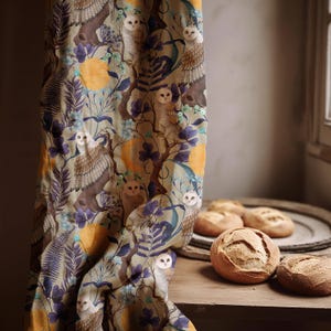May include: A curtain with a whimsical owl and floral print in shades of brown, purple, and blue. The fabric drapes over a wooden surface where several round loaves of bread are displayed. The curtain's design includes owls, trees, and foliage.
