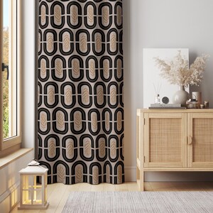 May include: A beige curtain with a repeating pattern of black ovals and tan leaf-like shapes. The curtain hangs in front of a window and next to a light-colored wooden cabinet with decorative items on top. A white lantern sits on the floor.